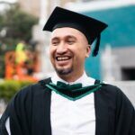 Male student in graduation cap and gown outside smiling