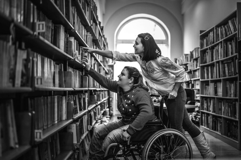 Support worker helping wheelchair user reach book on high shelf in university library