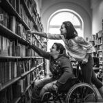 Support worker helping wheelchair user reach book on high shelf in university library